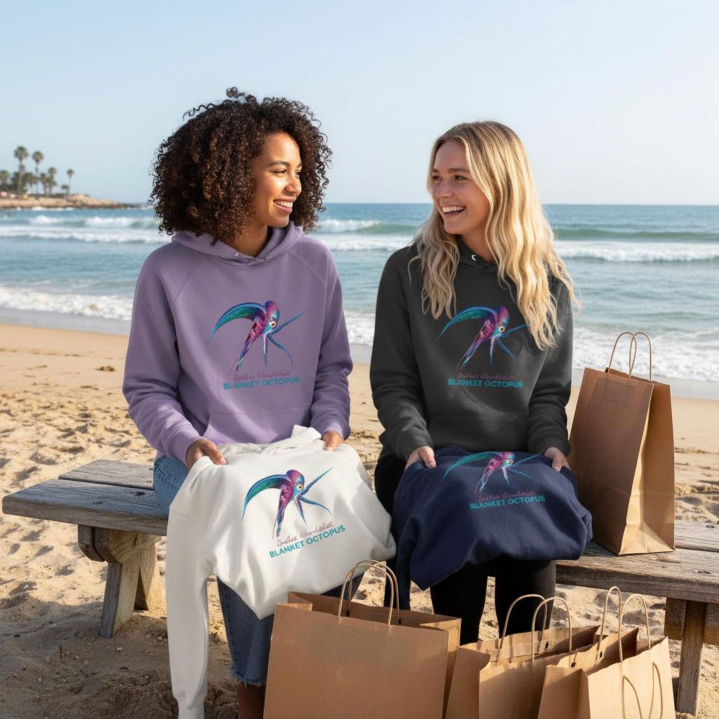 Two friends wearing organic Hoodies with the Blanket Octopus design by seablub in different colors, sitting on a bench by the ocean